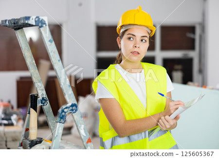 Woman foreman in a protective helmet and yellow vest checks the completed construction work on drawing Woman foreman in a protective helmet and yellow vest checks the completed construction work on drawing 125605367