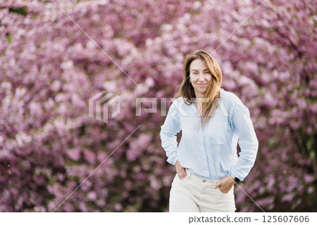Smiling woman standing among blooming pink cherry blossom sakura trees wearing a light blue shirt and enjoying a bright spring day outdoors 125607606