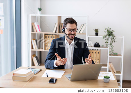 Young adult male in suit and headset smiling during online meeting, working at desk in bright office, expressing professional engagement and effective communication. Young adult male in suit and headset smiling during online meeting, working at desk in bright office, expressing professional engagement and effective communication. 125608108
