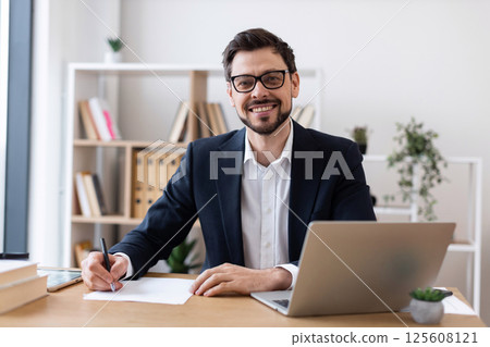 Smiling young Caucasian adult male in formal suit working at desk with laptop and papers. Professional, productive, and focused business setting showcasing modern office atmosphere. 125608121