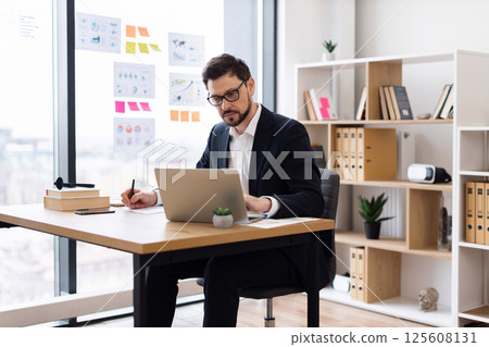 Young adult male wearing formal attire working with laptop at office desk writing notes. Modern workspace includes documents, books and potted plants indicating productive and professional atmosphere Young adult male wearing formal attire working with laptop at office desk writing notes. Modern workspace includes documents, books and potted plants indicating productive and professional atmosphere 125608131