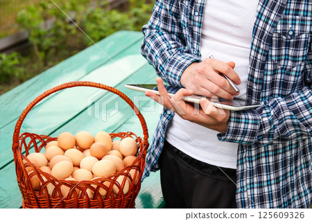 Man holding a tablet while inspecting eggs in a basket, representing quality control, food safety, and modern agricultural technology Man holding a tablet while inspecting eggs in a basket, representing quality control, food safety, and modern agricultural technology 125609326