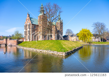 Rosenborg Castle stands majestically on its island surrounded by calm waters in Copenhagen. The clear blue sky enhances the castle's historic architecture, creating a picturesque view. 125611163