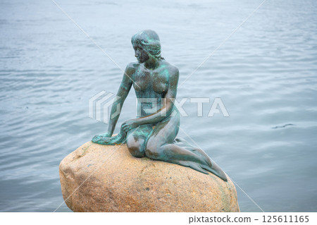 A bronze statue of a mermaid sits gracefully on a rock by the water in Copenhagen. Visitors admire the iconic sculpture, which is a symbol of the city and inspired by a tale. 125611165