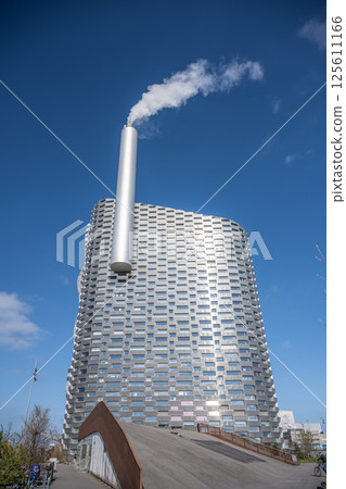 This modern facility combines waste-to-energy production with recreational activities. The unique architecture features a prominent smokestack, set against a clear blue sky in Copenhagen. 125611166