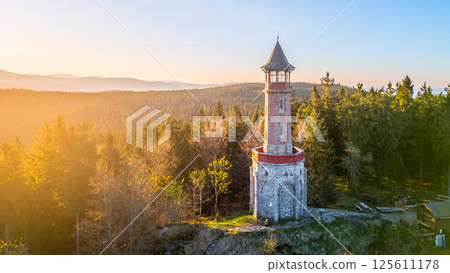 Stepanka Lookout Tower stands tall in the Jizera Mountains during a serene spring morning. The surrounding trees bask in warm sunlight, creating a tranquil natural setting. 125611178