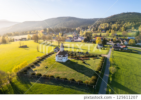 Tesarov Chapel, a striking white octagonal structure, stands amidst lush greenery in Korenov, surrounded by the picturesque Jizera Mountains. The serene landscape basks in soft sunlight. 125611207