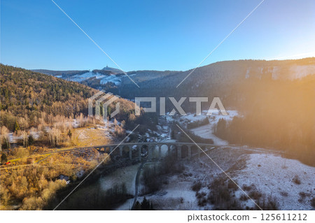 The railway stone bridge in Novina spans a tranquil valley, framed by snow-capped hills and Jested Mountain in the background during a beautiful winter sunset. 125611212