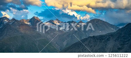 In the evening, the majestic alpine peaks are illuminated by soft light, casting dramatic shadows and highlighting the rugged terrain. Clouds swirl overhead, adding depth to the stunning summer 125611222