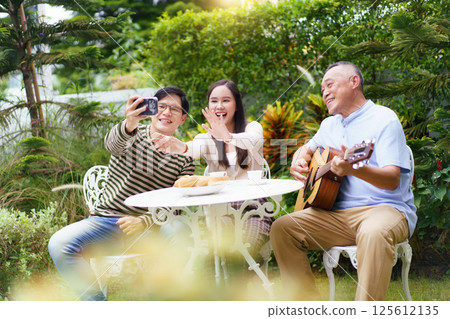 Happy Asian family spending time together, man and woman let their father playing a guitar and singing in backyard garden at home. Mental health care and wellbeing in senior adult. 125612135