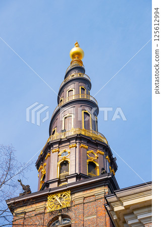 The corkscrew spire of Church of Our Saviour towers majestically over the Copenhagen skyline against a clear blue sky. This iconic structure showcases its intricate design and golden top. 125612994