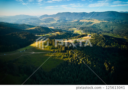 Aerial view of picturesque valley with rolling hills, lush forests, and scattered houses. Sunlight bathes landscape in warm glow, highlighting vibrant greens and creating serene atmosphere. 125613041