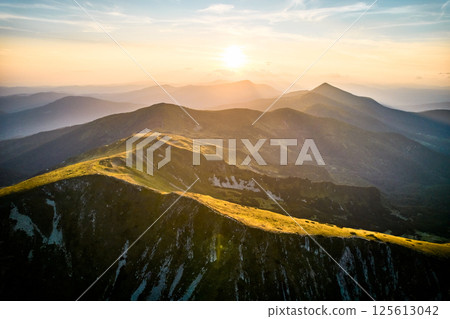 Aerial view of rugged mountain range with lush green slopes at sunset. Landscape showcases beauty of untouched nature with distant peaks and valleys bathed in soft sunlight. Carpathians, Chornogora. 125613042