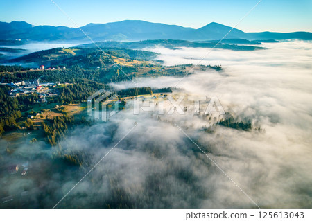 Aerial view of lush, green valley with scattered houses bathed in early morning sunlight. Mist rolls through forested hills, creating ethereal atmosphere against backdrop of distant mountains. Aerial view of lush, green valley with scattered houses bathed in early morning sunlight. Mist rolls through forested hills, creating ethereal atmosphere against backdrop of distant mountains. 125613043