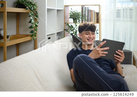 Smiling man relaxing on sofa watching content on digital tablet with wireless earbuds Smiling man relaxing on sofa watching content on digital tablet with wireless earbuds 125614249