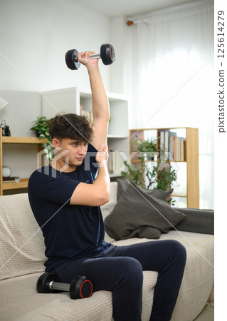 Young man exercising with dumbbells on sofa. Healthy lifestyle and home workouts 125614279