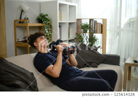 Young man exercising with dumbbells on sofa. Healthy lifestyle and home workouts 125614326