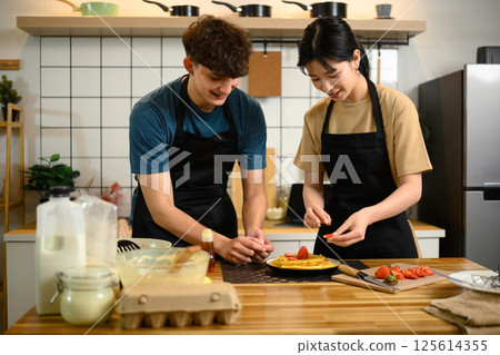 Happy young couple decorating homemade pancakes with fresh strawberries in a kitchen 125614355