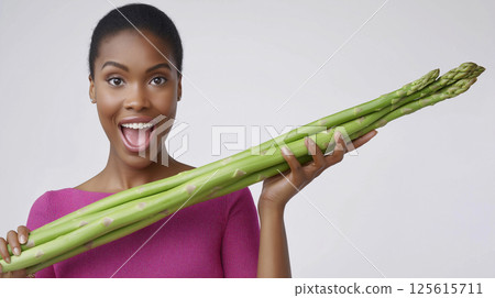 An adult woman smiles widely while proudly displaying a large bunch of asparagus. She stands against a light background, celebrating fresh vegetables and healthy living. 125615711