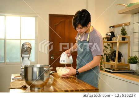 Young asian man using a hand mixer to churn cream in a glass bowl Young asian man using a hand mixer to churn cream in a glass bowl 125616227
