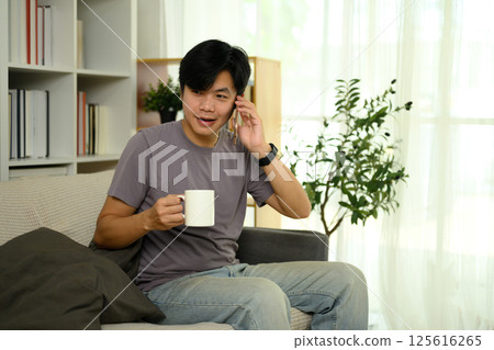 Smiling asian man holding a cup of coffee and talking on mobile phone in a living room 125616265
