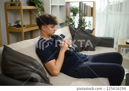 Tired young caucasian male sitting on sofa with dumbbell in living room 125616299