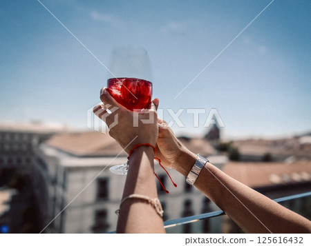 A glass of red wine or tinto de verano in a woman's hand against a blue sky and cityscape. People at a party celebrating on a restaurant terrace.  125616432