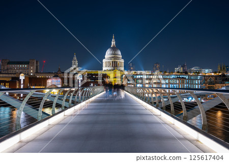 The Millennium Bridge spans the River Thames, illuminated under the evening sky. St. Paul's Cathedral stands majestically in the background, surrounded by city lights. 125617404