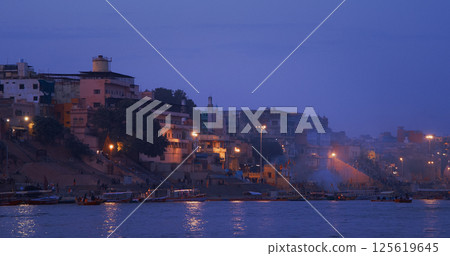 Varanasi, Uttar Pradesh, India. People walking on steps on Shivala Ghat and floating in boats near Dandi Ghat. Evening time. Camera moving along riverbank embankment. Cinematic View from river 125619645