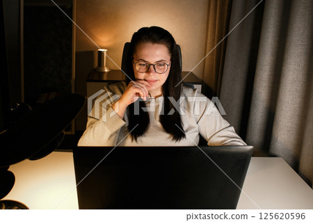 Smiling millennial girl sitting at desk with computer scrolling internet, doing online shopping. Working on laptop in home office. Freelance work at home. Smiling millennial girl sitting at desk with computer scrolling internet, doing online shopping. Working on laptop in home office. Freelance work at home. 125620596