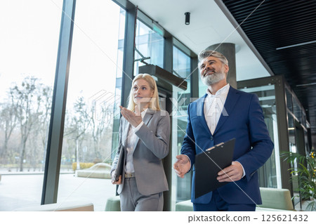 Manager and employee going through corridor holding laptop discussing teamwork 125621432