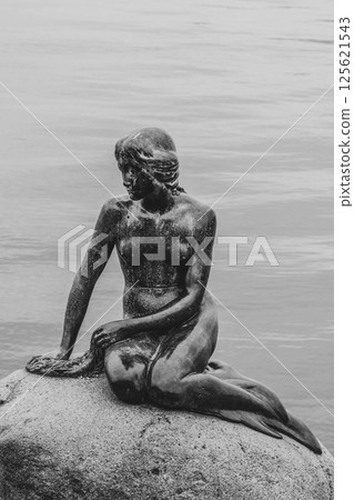 The Little Mermaid statue showcases its enchanting beauty while perched on a rock by the waterfront in Copenhagen, Denmark, inviting visitors to admire its charm against the serene backdrop. 125621543