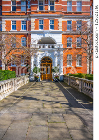 The red brick building features elegant architecture with a grand entrance surrounded by manicured hedges. Bright sunlight illuminates the path leading to the entrance, enhancing the vibrant details. The red brick building features elegant architecture with a grand entrance surrounded by manicured hedges. Bright sunlight illuminates the path leading to the entrance, enhancing the vibrant details. 125621566