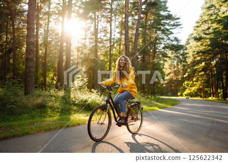Young happy woman in yellow coat rides bicycle in sunny park. Woman enjoys autumn nature. Lifestyle. 125622342