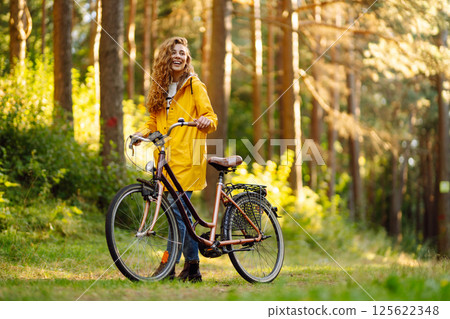 Young happy woman in yellow coat rides bicycle in sunny park. Woman enjoys autumn nature. Lifestyle. 125622348