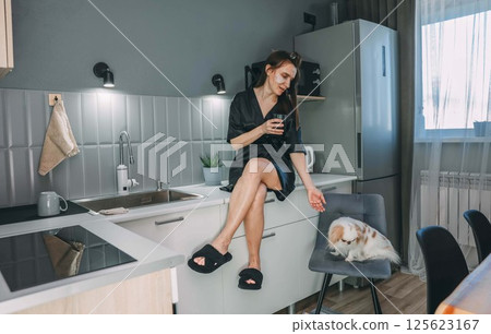 relaxed woman enjoying a glass of water in modern kitchen, with small dog, concept of daily hydration 125623167