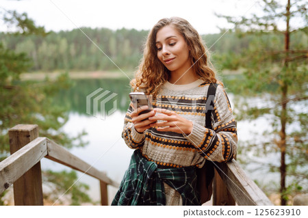 A young woman in a cozy sweater walking near a beautiful lake, smiling and using her phone 125623910