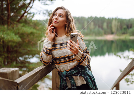 A young woman in a cozy sweater walking near a beautiful lake, smiling and using her phone 125623911