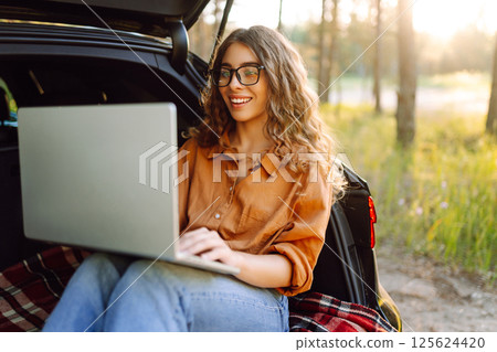 Young woman using laptop sitting in car trunk during sunset. Working while on vacation by car. Young woman using laptop sitting in car trunk during sunset. Working while on vacation by car. 125624420