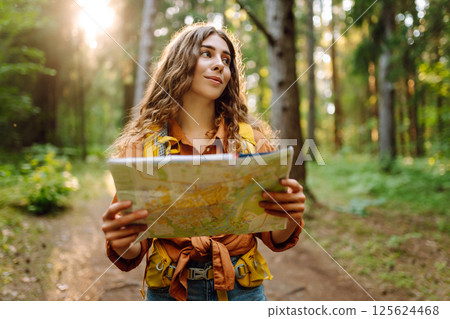 A traveler woman stands in a lush forest, examining a map with a focused expression 125624468