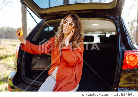 Happy woman sits on the trunk of her car and taking a selfie. Selfie while traveling by car. Happy woman sits on the trunk of her car and taking a selfie. Selfie while traveling by car. 125624643