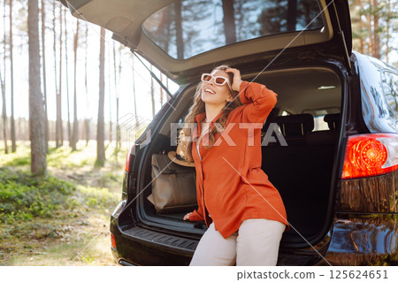 A traveler young woman smiles while sitting in the trunk of her car during a sunny day in a forest 125624651