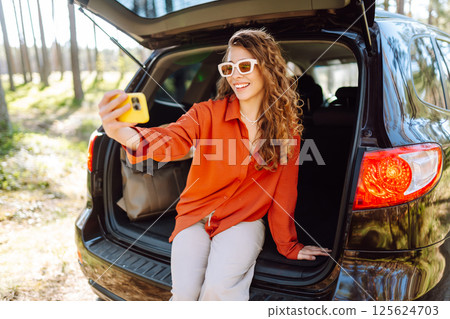 Happy woman sits on the trunk of her car and taking a selfie. Selfie while traveling by car. Happy woman sits on the trunk of her car and taking a selfie. Selfie while traveling by car. 125624703