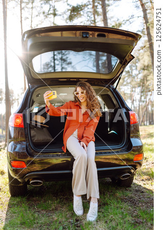 Happy woman sits on the trunk of her car and taking a selfie. Selfie while traveling by car. Happy woman sits on the trunk of her car and taking a selfie. Selfie while traveling by car. 125624751
