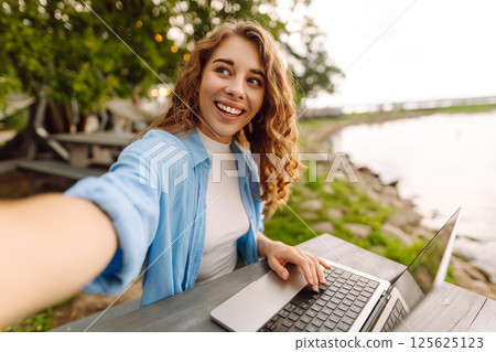 Happy woman in stylish clothes sitting at a table outdoors taking a selfie, working on a laptop. 125625123
