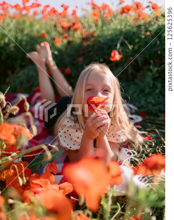 Blonde child girl on a plaid in blooming poppy field, summer day 125625396