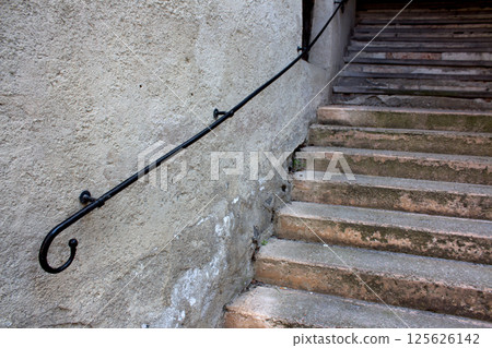 A weathered stone staircase ascends alongside a rough, textured wall, guided by a simple black metal handrail. The steps show signs of age and use 125626142