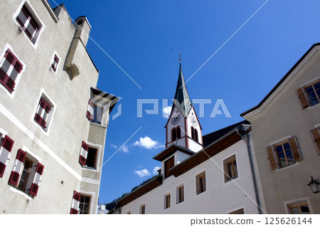 A charming upward view of the steeple of St. Virgil Parish Church in Rattenberg, Austria. The steeple, with its clock face, stands tall against a bright blue sky dotted with a few white clouds A charming upward view of the steeple of St. Virgil Parish Church in Rattenberg, Austria. The steeple, with its clock face, stands tall against a bright blue sky dotted with a few white clouds 125626144