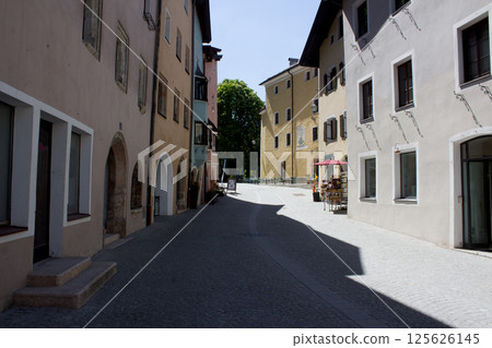 A quiet Rattenberg street lined with colorful buildings under a bright sky, casting shadows along the pavement towards a distant green tree 125626145