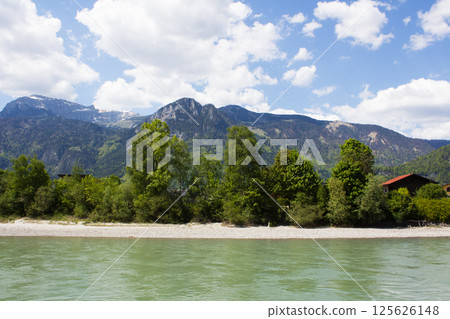 A serene turquoise river flows in the foreground, bordered by a light rocky shore and lush green trees, with majestic mountains rising against a partly cloudy blue sky A serene turquoise river flows in the foreground, bordered by a light rocky shore and lush green trees, with majestic mountains rising against a partly cloudy blue sky 125626148